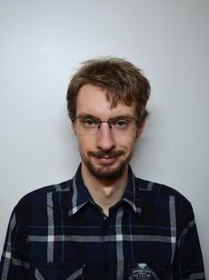 Junger Mann mit kariertem dunklem Hemd vor grauem Hintergrund; Gesicht durch Rechteck verdeckt.
